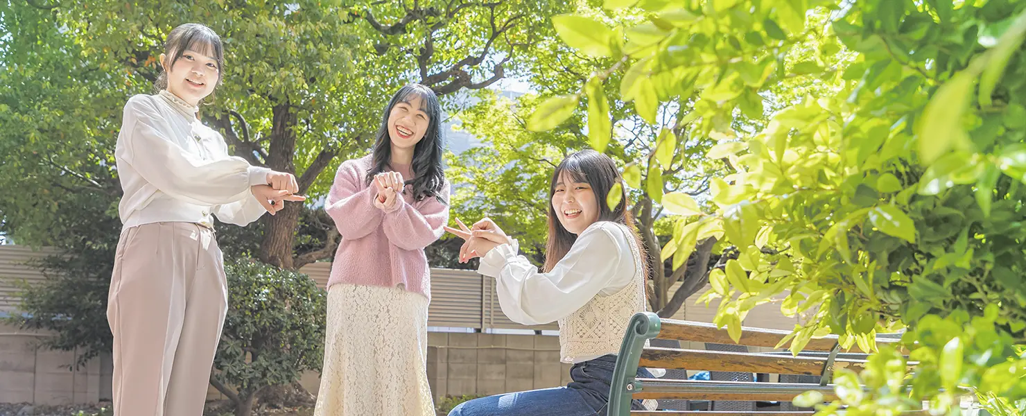 屋外の緑に囲まれたベンチのそばで、3人の女子学生が笑顔でポーズを取っている。明るく和やかな雰囲気の中、学生同士の交流の様子が伝わる。