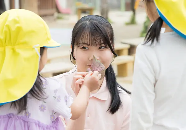 保育実習中の学生が、園児と葉っぱを使って楽しくふれあう様子。笑顔があふれる関わりのひとコマ。